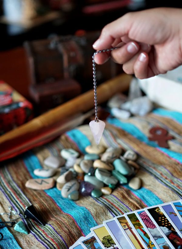 A hand holds a pendulum over a collection of tarot cards and stones, suggesting a mystical or spiritual setting.